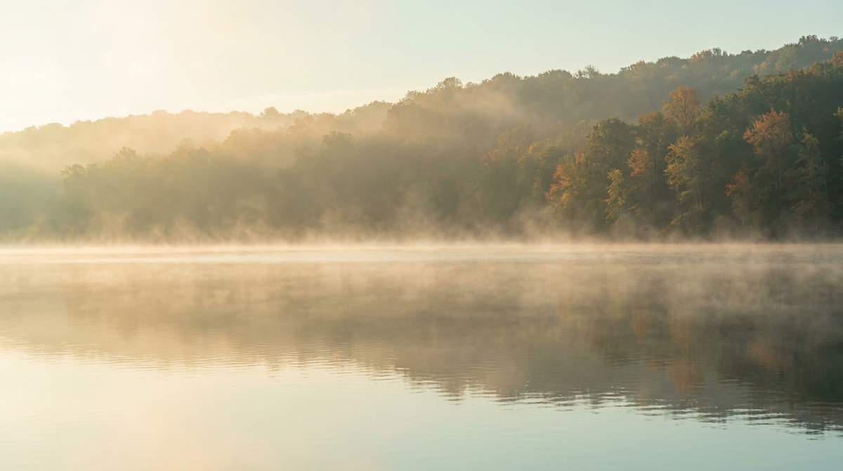A calm freshwater reservoir at early autumn dawn with low mist drifting across a glassy surface and a distant treeline beginning to turn color.
